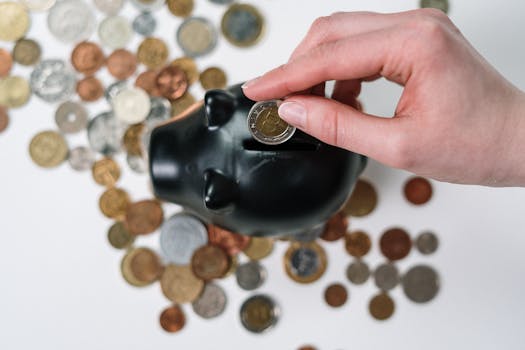 Close-up of a hand inserting a coin into a black piggy bank with scattered coins on a white background.